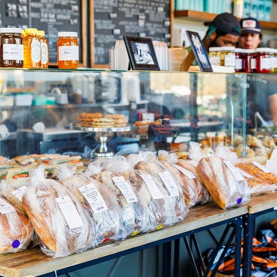 Deli counter displaying fresh bread, sandwiches, pastries, and jars of honey, with staff working.